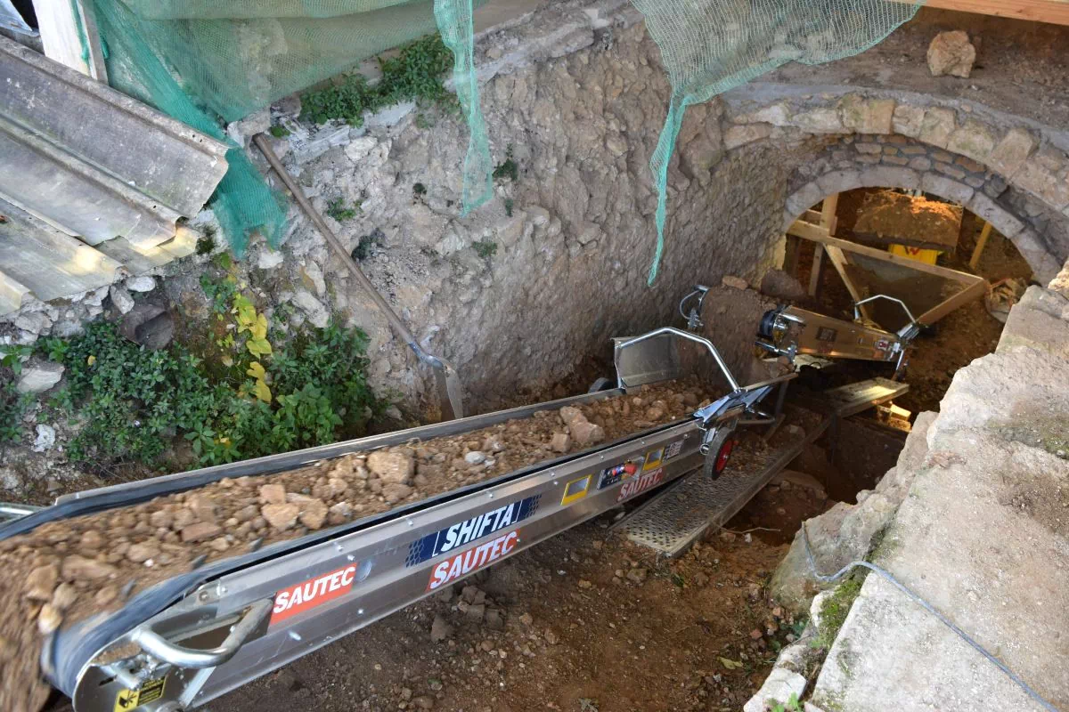 Un mini-dumper déverse de la terre dans la trémie d'un tapis convoyeur.
